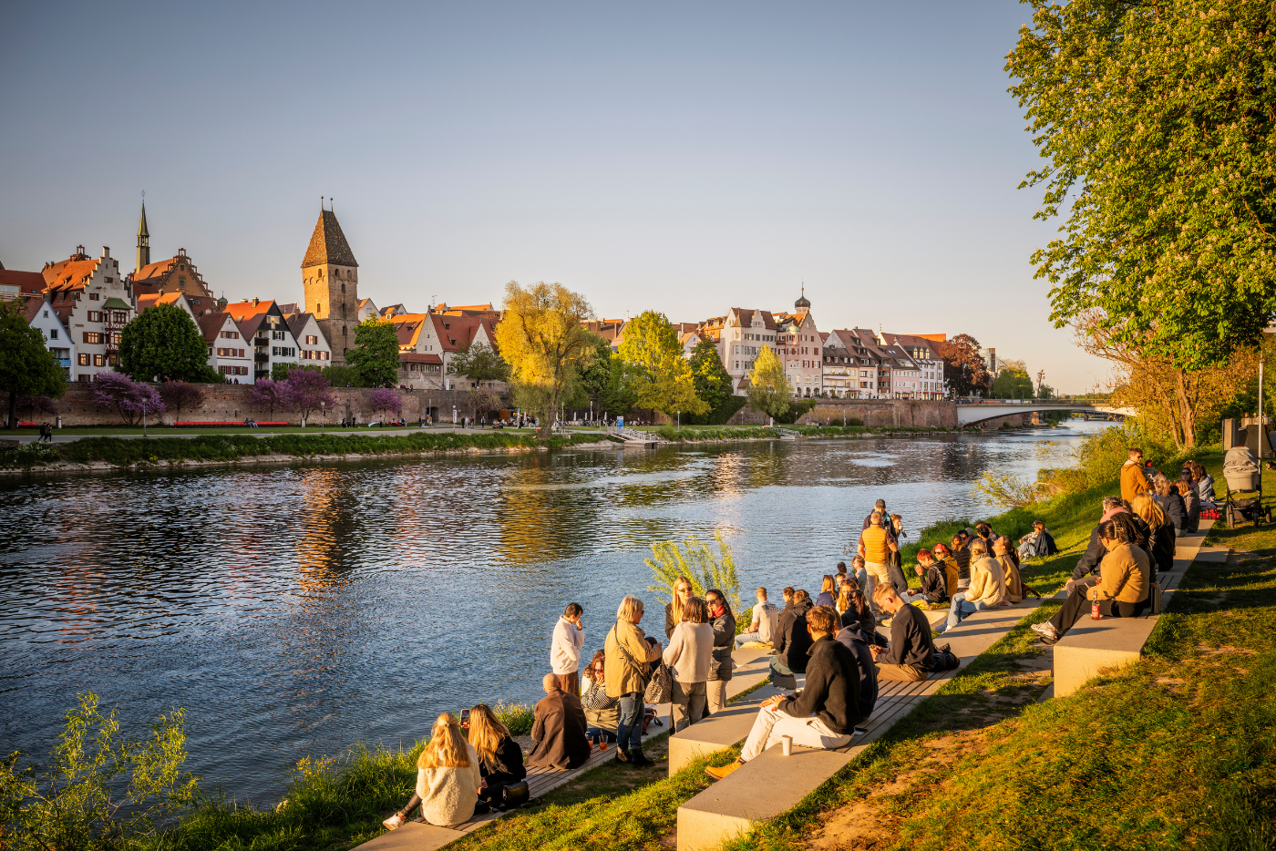 Mensen zitten aan de oever van de Donau in Ulm, met de historische binnenstad op de achtergrond.