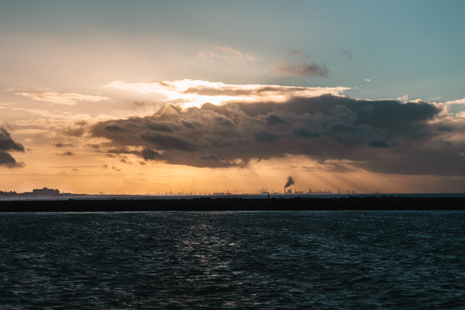 Zonlicht breekt door donkere wolken boven het water, met een silhouet van de horizon op de achtergrond.