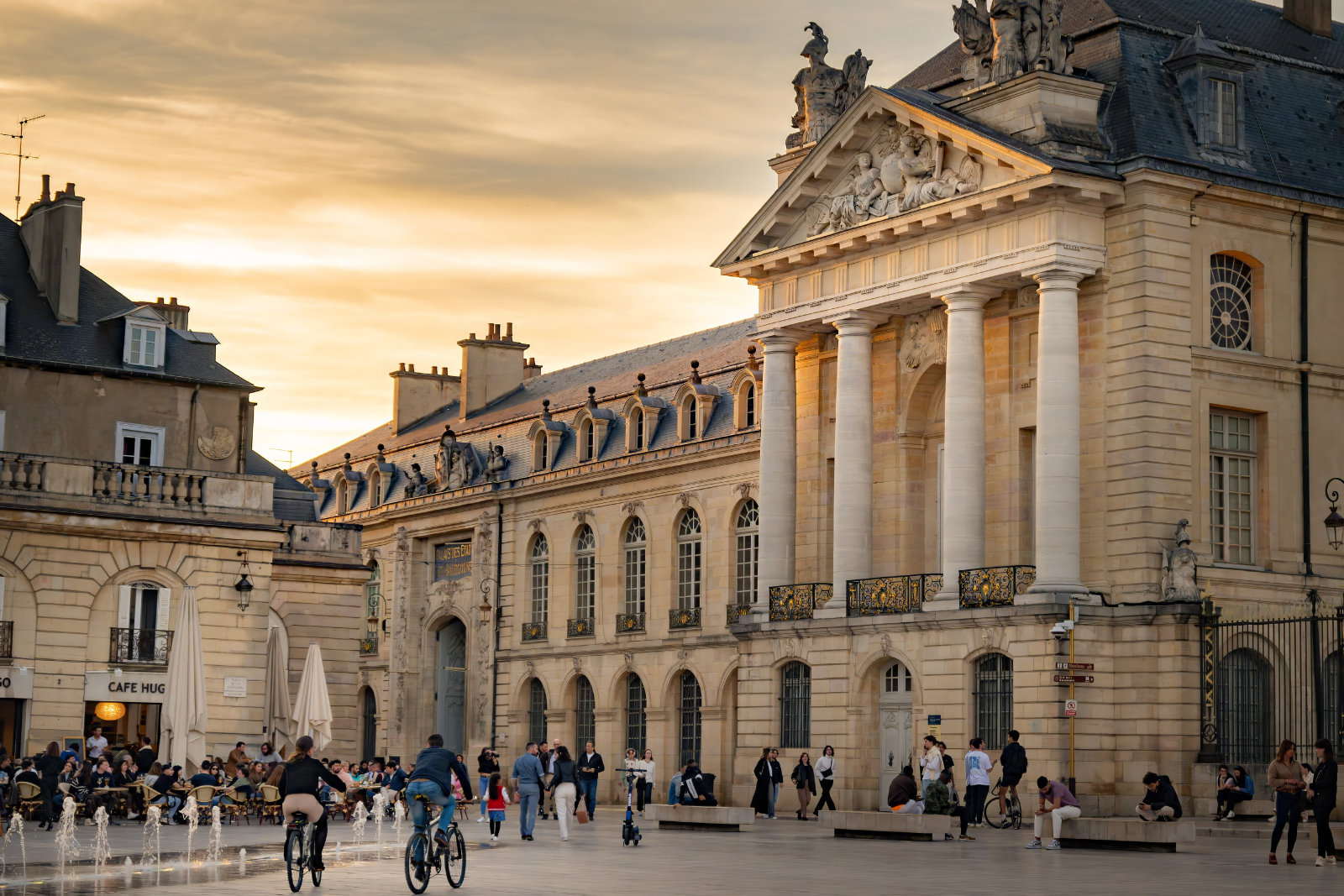 Fietsers rijden over een levendig plein in Dijon met terrassen en historische gebouwen op de achtergrond.
