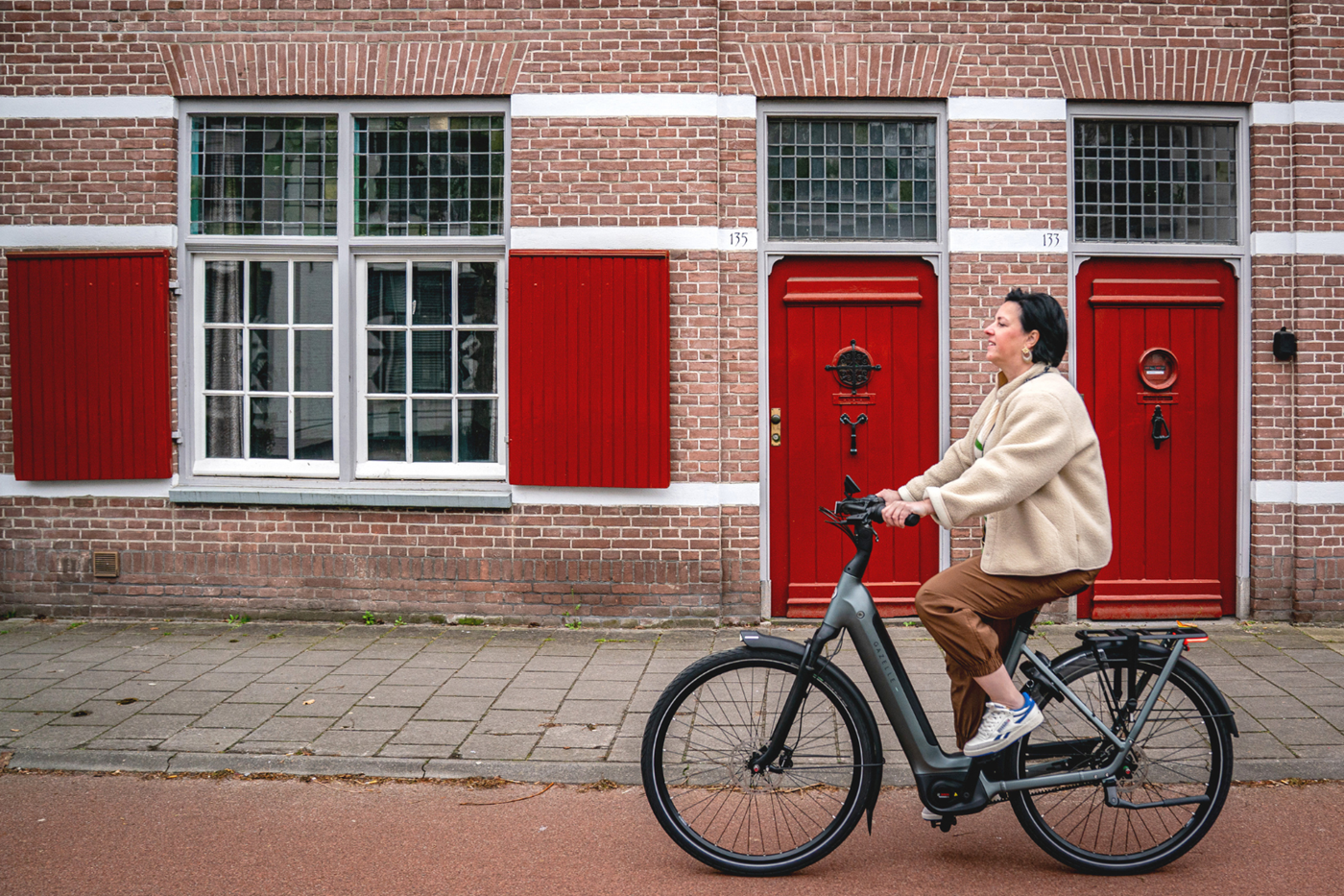 Vrouw fietst op een elektrische fiets langs een rij historische woningen met rode deuren.