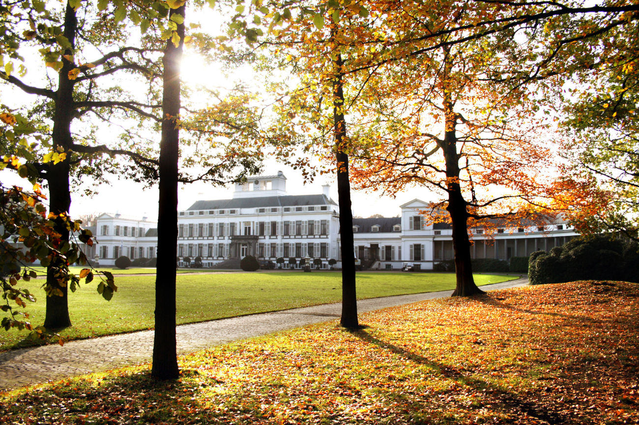 Paleis Soestdijk tussen bomen met herfstkleuren en afgevallen bladeren.