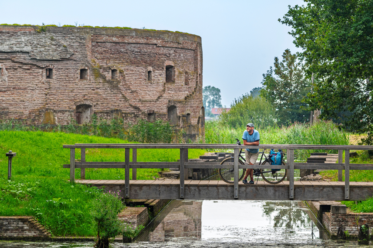 Man met fiets leunt op een houten brug bij een historisch fort, omringd door water en groen.