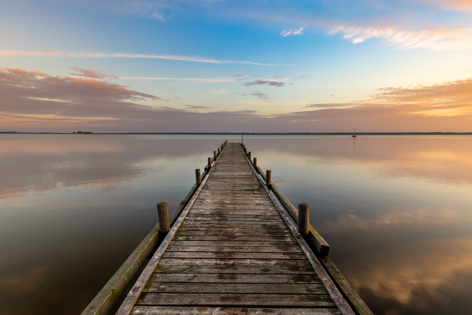 Houten steiger aan het Steinhuder Meer tijdens zonsondergang, met rustig water en een zeilboot in de verte.